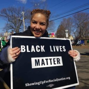 Felicia, wearing a warm coat and holding a Black Lives Matter sign in front of a blue sky